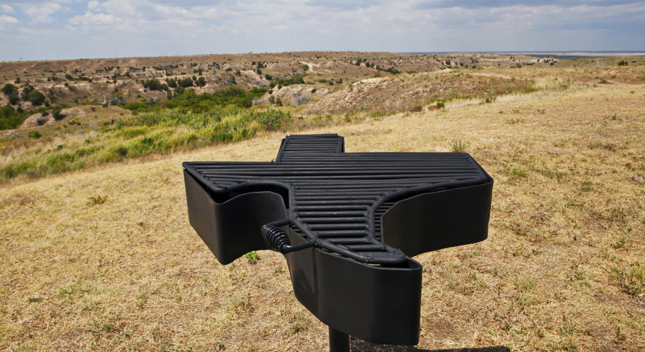 A Texas-shaped barbeque grill with rugged Texas scenery in the background A Texas-shaped barbeque grill with rugged Texas scenery in the background